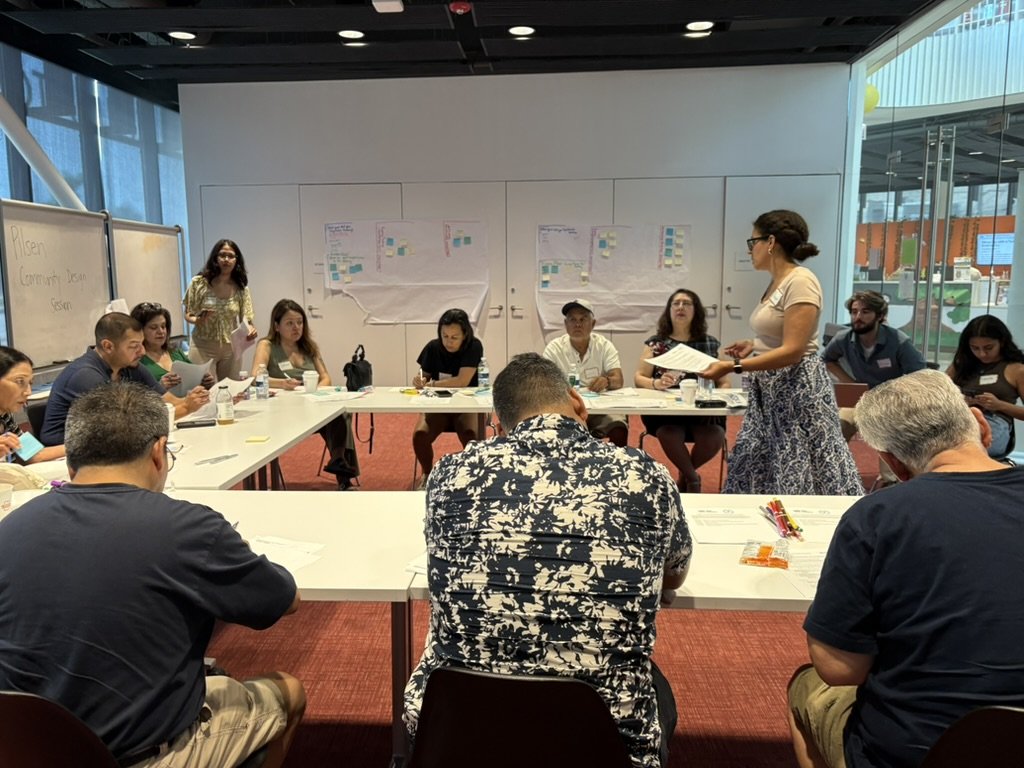 A group meeting in a conference room with a speaker standing and others seated around tables.